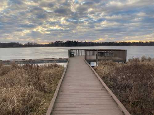 A wooden walkway leads to a dock over a calm lake, with cloudy skies and trees in the background.