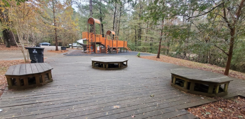 A wooden deck with benches, surrounded by trees, featuring a playground structure in the background.