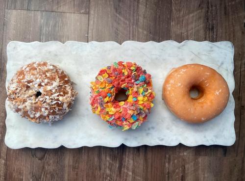 Three colorful donuts on a marble platter: one with coconut, one with fruity cereal, and a plain glazed donut.