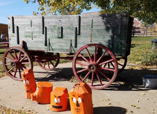 A wooden wagon with red wheels beside three orange pumpkin-shaped decorations with faces, set in a sunny outdoor area.