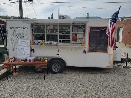 A food truck with a menu board, decorated with lights and an American flag, parked on gravel with a wooden table of treats.