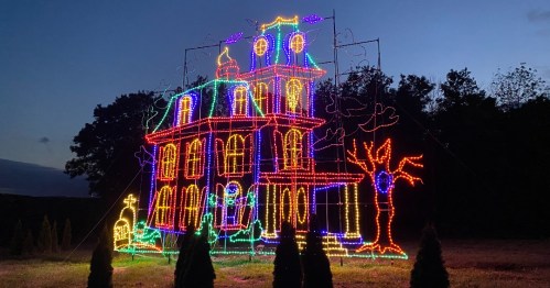 A brightly lit, colorful haunted house made of lights, set against a twilight sky with trees in the background.