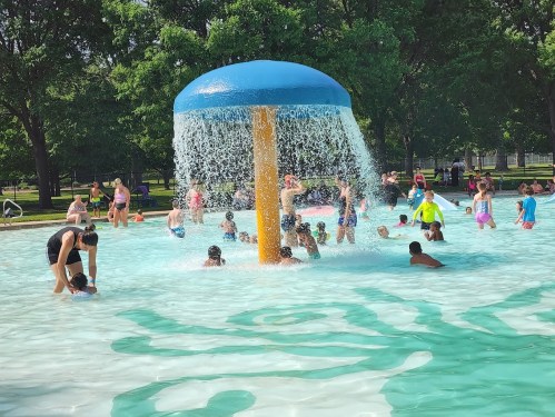 A vibrant water park scene with children playing under a large blue mushroom fountain in a sunny outdoor pool.