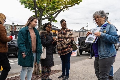 A group of four women stands outdoors, listening to an older woman showing a photo in a booklet.