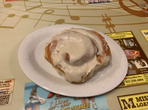 A cinnamon roll with cream cheese frosting on a white plate, resting on a table with colorful advertisements.