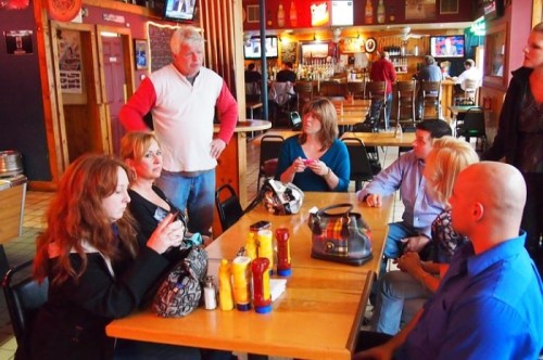 A group of people sitting at a table in a casual restaurant, engaged in conversation with a man standing nearby.
