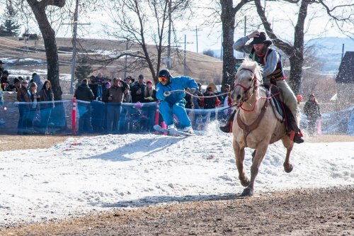 A cowboy on a horse races alongside a skier, with a crowd watching in a snowy landscape.