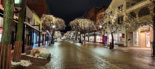 A quiet street at night, lined with trees adorned with twinkling lights and snow on the ground.
