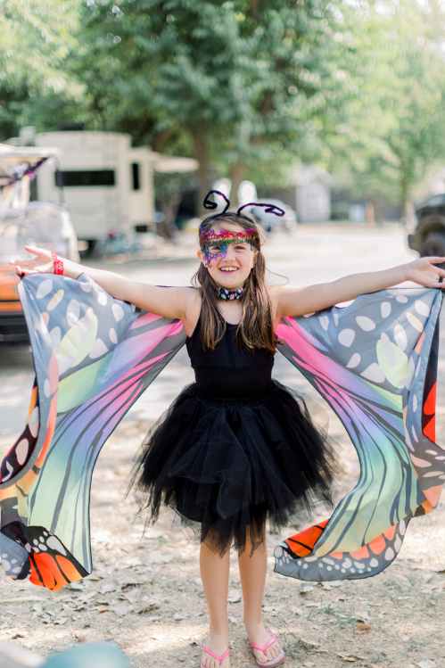 A young girl in a butterfly costume with colorful wings and a black tutu, smiling and posing outdoors.