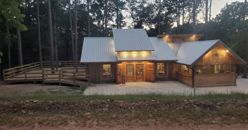 A rustic wooden house with a metal roof, surrounded by trees, featuring warm lights and a spacious deck.