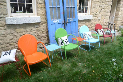 Colorful chairs in a row outside a stone building with blue doors, surrounded by green grass.