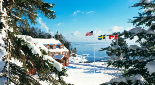 A snowy landscape featuring a red building, flags of the US, Sweden, and Canada, with a serene lake in the background.