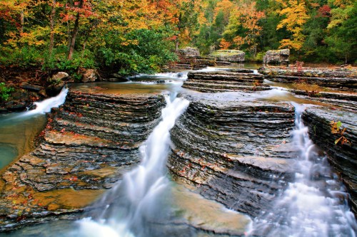 A serene waterfall cascades over layered rocks, surrounded by vibrant autumn foliage in a tranquil forest setting.