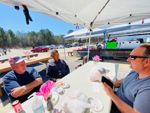 Three men sit at a table under a tent, enjoying food and drinks, with a sunny outdoor setting and trees in the background.