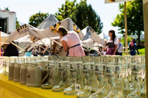 A row of glass beer boots and mugs on a table, with people in traditional attire in the background at an outdoor festival.