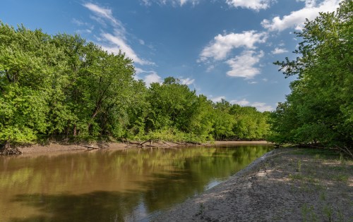 A serene riverbank surrounded by lush green trees under a partly cloudy sky.