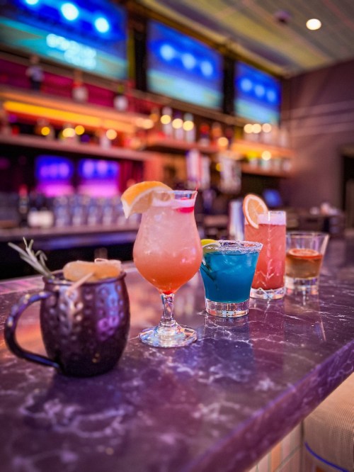 A vibrant bar scene featuring four colorful cocktails on a marble countertop, with a blurred background of bar shelves.