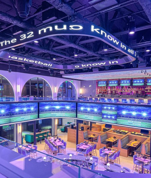 A modern sports bar with multiple screens, vibrant lighting, and seating areas, featuring a large illuminated sign overhead.