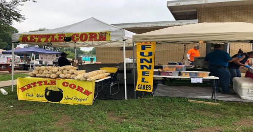 A food stall featuring kettle corn and funnel cakes at an outdoor event, with colorful signage and people in the background.