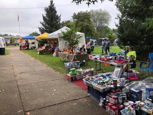 A vibrant outdoor market with various vendor tents, tables of goods, and people browsing in a green park setting.