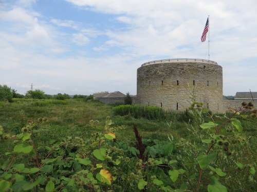 A round stone fort with an American flag, surrounded by green grass and plants under a cloudy sky.