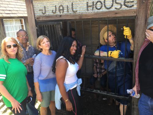 A group of people poses near a jailhouse prop, with one person inside wearing a straw hat and gloves.