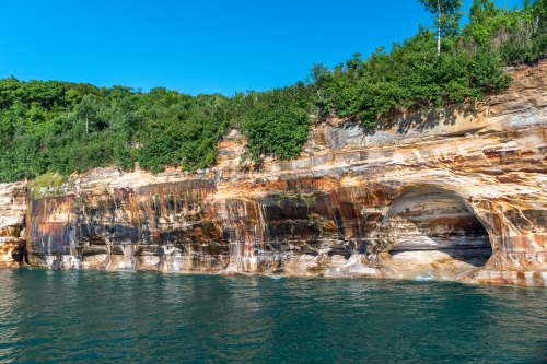 Colorful rock formations and a natural arch along a shoreline, with clear blue water and green foliage above.