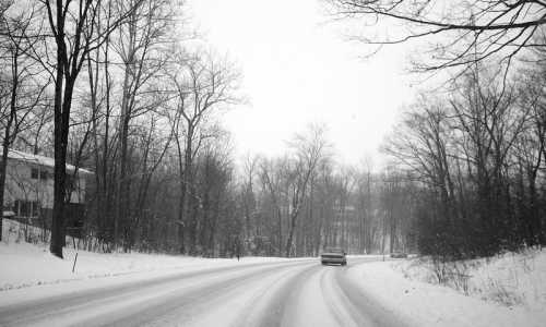 A snowy road winds through a forest, with trees bare of leaves and a car driving in the winter weather.