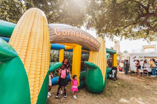Children play near a colorful inflatable corn maze at an outdoor event, surrounded by trees and festive decorations.