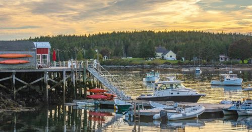A serene harbor at sunset, featuring boats docked by a wooden pier and a backdrop of lush green trees.