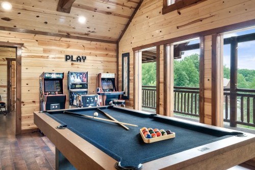 A cozy game room with a pool table, arcade machines, and large windows overlooking greenery.