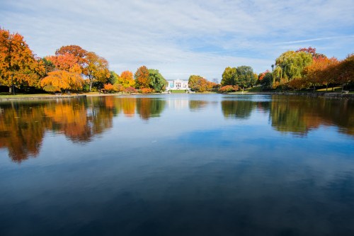 A serene lake reflects colorful autumn trees and a white building under a blue sky with scattered clouds.