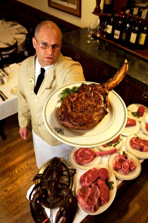 A waiter in a beige jacket holds a large steak on a plate, surrounded by various cuts of meat and a lobster.
