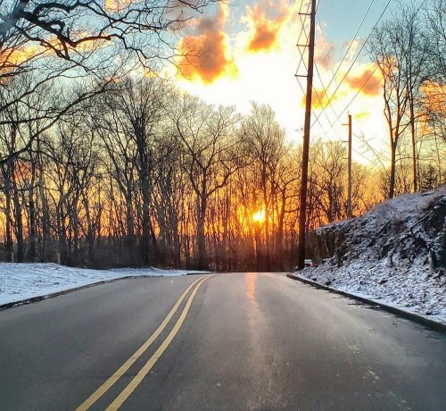 A winding road through a snowy landscape, with a vibrant sunset and silhouetted trees in the background.