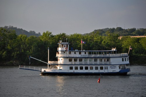 A vintage riverboat sails on a calm waterway, surrounded by lush green trees and a distant shoreline.