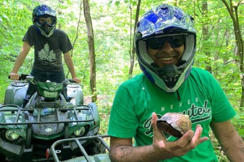 Two people in helmets on an ATV in a forest, one holding a turtle and smiling.