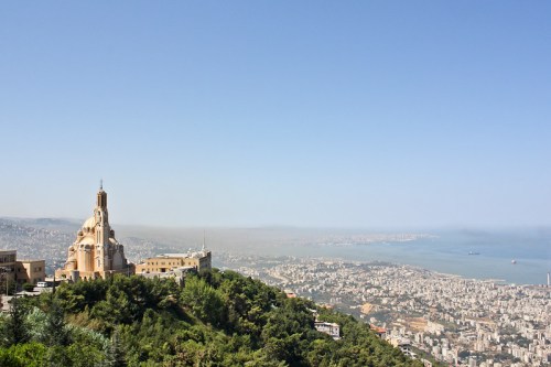 A panoramic view of a coastal city with a prominent church on a hill, surrounded by greenery and blue skies.