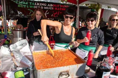 A woman stirs a large pot of chili at a festival, surrounded by friends, all wearing black tops and green aprons.