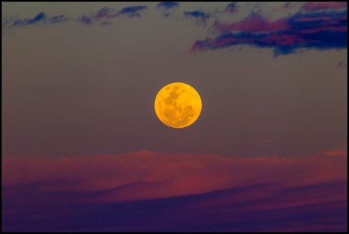 A large, bright orange moon rises against a colorful twilight sky with soft clouds.