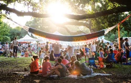 A lively outdoor gathering with people sitting on the grass, enjoying a sunny day under a large tree and colorful decorations.