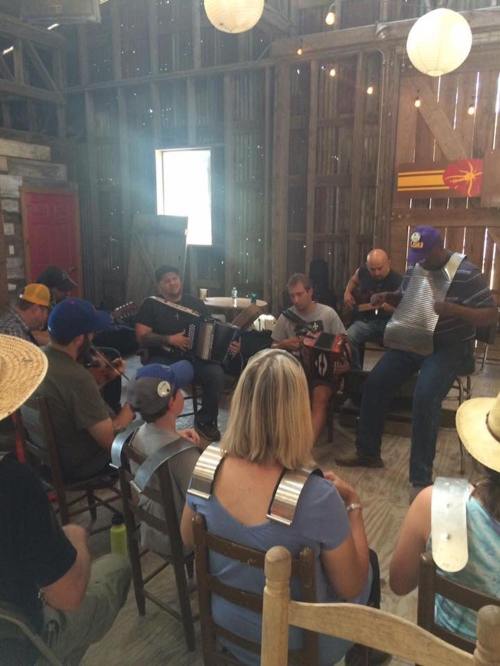 A group of musicians playing instruments in a rustic barn setting, with seated audience members enjoying the performance.