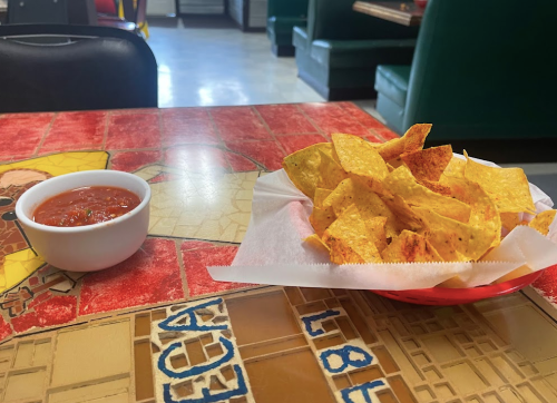 A basket of tortilla chips next to a small bowl of salsa on a colorful table in a restaurant.