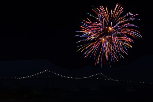 Colorful fireworks burst in the night sky above a bridge, creating a vibrant display against a dark backdrop.