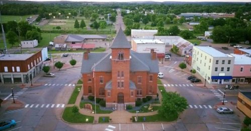 Aerial view of a historic brick building in a small town, surrounded by trees and shops, with a clear sky above.