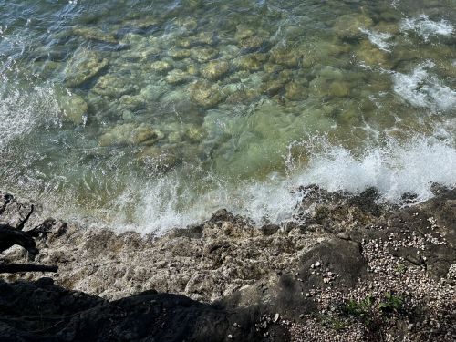Waves gently crash against rocky shore, revealing clear water and visible stones beneath the surface.