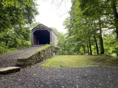 A covered bridge surrounded by lush green trees and a gravel path, with a person standing inside the bridge.