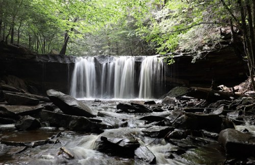 A serene waterfall cascades over rocks, surrounded by lush green trees in a tranquil forest setting.
