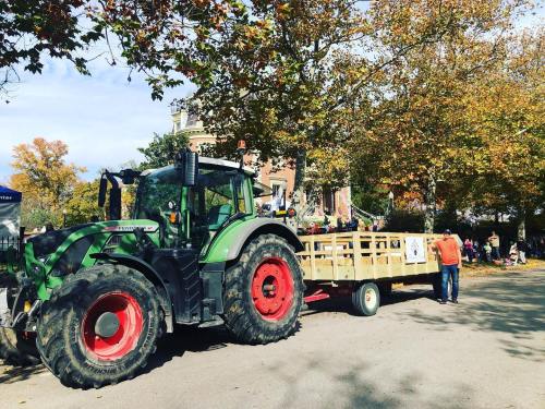 A green tractor with a wooden trailer parked on a street lined with trees and people in the background.