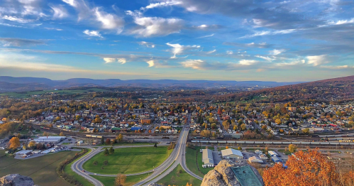 A panoramic view of a small town surrounded by hills, with roads and autumn foliage under a blue sky.