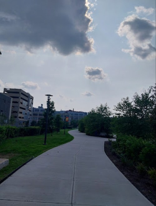 A winding pathway through a green park, with buildings in the background and a cloudy sky above.